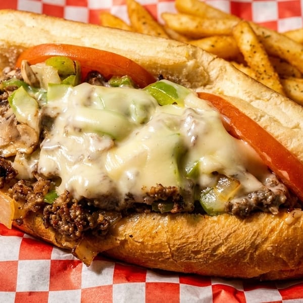 Cheesesteak sandwich with melted cheese, peppers, onions, and tomatoes, served with fries on checkered paper.